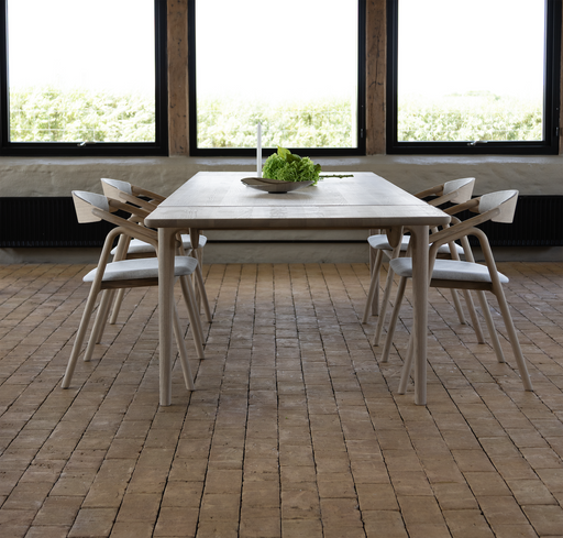 A Floating Dining Table with four wooden chairs stands on a brick floor before three large windows, its center adorned by a shallow bowl filled with green leaves.