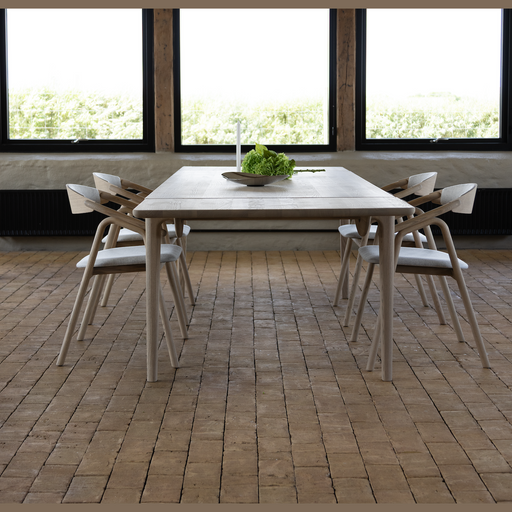 A Floating Dining Table with four wooden chairs stands on a brick floor before three large windows, its center adorned by a shallow bowl filled with green leaves.
