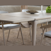 A close-up of the Floating Dining Table in ash wood with a partially extended leaf, surrounded by matching chairs with light grey cushions on a tiled floor. A bowl and greenery atop enhance its elegant floating effect.