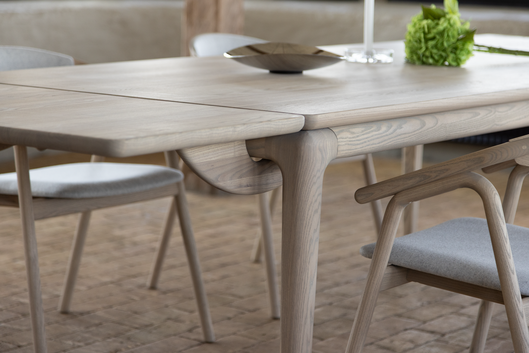 A close-up of the Floating Dining Table in ash wood with a partially extended leaf, surrounded by matching chairs with light grey cushions on a tiled floor. A bowl and greenery atop enhance its elegant floating effect.