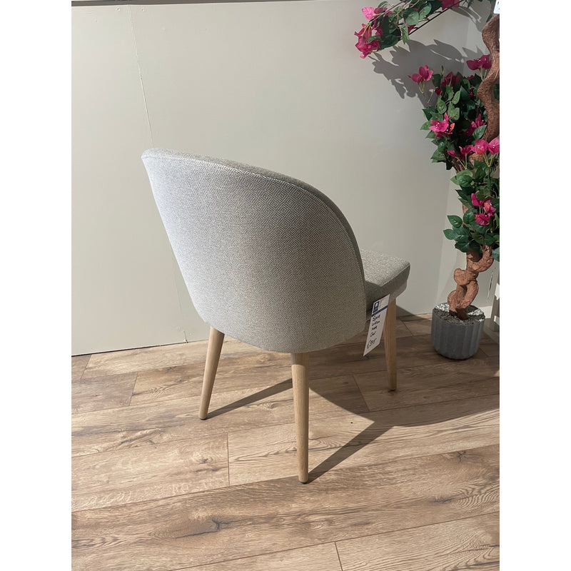 The Space dining chair, featuring light grey upholstery and wooden legs, sits on a wooden floor near a beige wall. Beside it is a potted plant with pink flowers, and a tag hangs from the chair’s leg.
