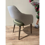 The Turim Dining Chair features a textured gray back, olive green cushioned seat, and wooden legs. Placed on a wooden floor beside a potted plant with pink flowers against a light-colored wall, it adds modern style to any space.