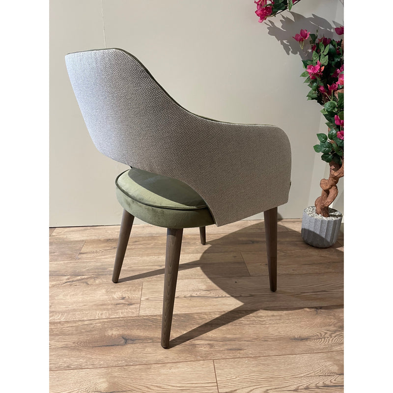 The Turim Dining Chair features a textured gray back, olive green cushioned seat, and wooden legs. Placed on a wooden floor beside a potted plant with pink flowers against a light-colored wall, it adds modern style to any space.