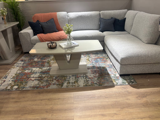 A modern living room featuring the Sorrento Chaise Corner in neutral fabric, dark and light pillows, an orange blanket, a glass-top coffee table, a colorful rug, wood flooring, and small vases with flowers on the table and sideboard.
