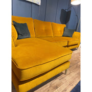 The Turino Chaise, a mustard yellow velvet corner sofa with dark green cushions in aquaclean fabric, rests on a wooden floor by a navy blue paneled wall, bathed in the light of a floor lamp.