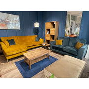 A modern living room showcases the Turino mustard yellow sectional sofa with aquaclean fabric by vendor-unknown, a teal blue Irish-made sofa, wooden coffee table, bookshelf, and blue walls. Accent pillows, floor lamp, and wall art finish the cozy look.