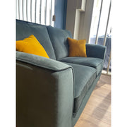 The Turino sofa by vendor-unknown, in dark green, features two mustard yellow cushions and sits on a wooden floor by a window with vertical blinds. Sunlight streams in, creating patterns of light and shadow on the sofa and floor.