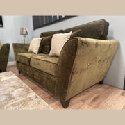 A Cosmos Sofa in green velvet with three beige and patterned throw pillows sits on a light wooden floor next to a beige rug, set against a light blue wall with a lamp in the background.