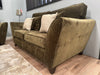 A Cosmos Sofa in green velvet with three beige and patterned throw pillows sits on a light wooden floor next to a beige rug, set against a light blue wall with a lamp in the background.