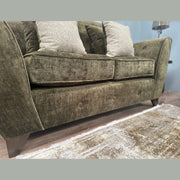 A close-up of the Cosmos Sofa in green velvet with patterned beige cushions on wooden legs, set on a light textured rug with a small lamp visible in the background.