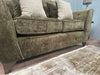 A close-up of the Cosmos Sofa in green velvet with patterned beige cushions on wooden legs, set on a light textured rug with a small lamp visible in the background.