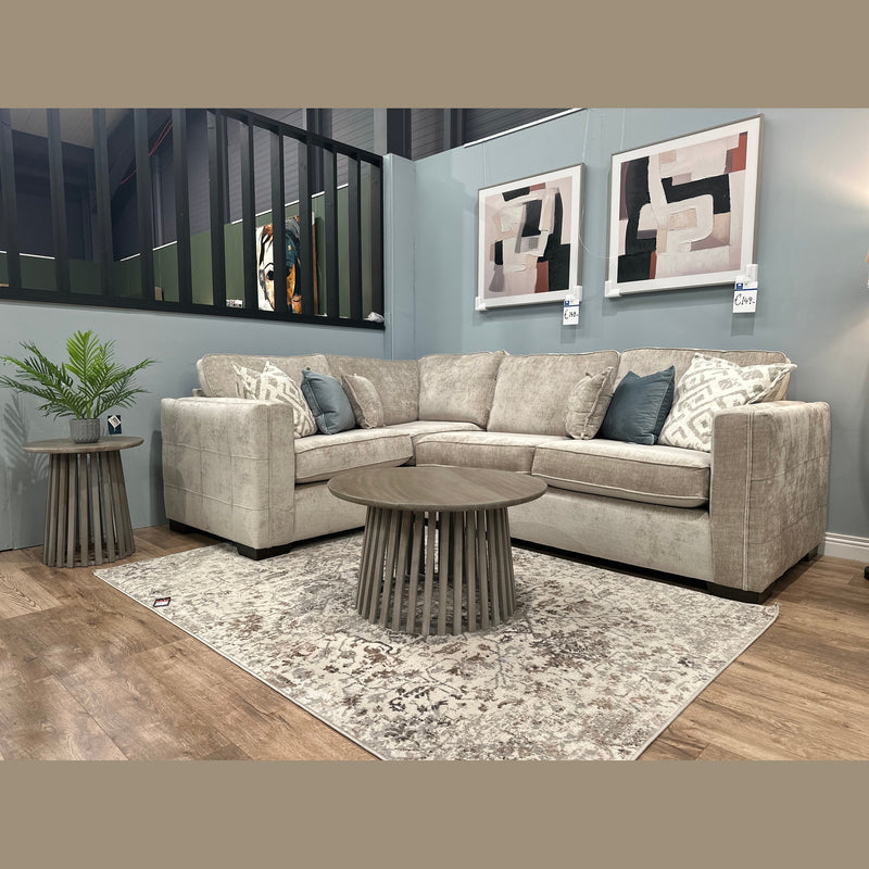 A cozy living room featuring the Ivy Corner Sofa in premium beige fabric, adorned with blue and patterned cushions, two round wooden tables, a potted plant, abstract wall art, and a patterned rug on wooden flooring.