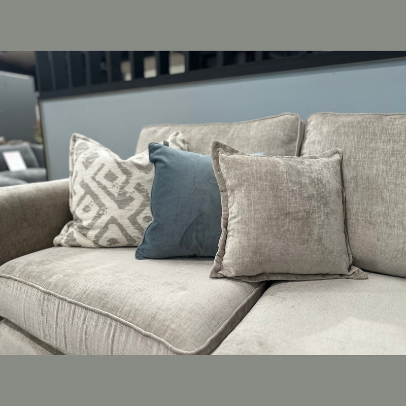 The Ivy Corner Sofa features a light gray, contemporary design with three neatly arranged back cushions: one geometric patterned, one solid blue, and one solid gray.