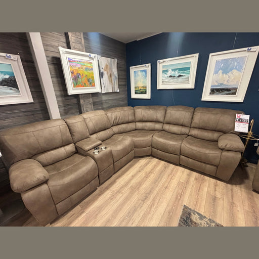 The Sapphire Corner Group, a spacious brown reclining sofa with cup holders, is displayed in a showroom with blue and wood-paneled walls, wooden flooring, and colorful landscape and seascape paintings above.