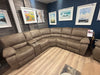 The Sapphire Corner Group, a spacious L-shaped brown recliner with cup holders, is displayed on wood flooring. Framed landscape art decorates the dark blue wall behind it, and a price tag is attached to one of the seats.