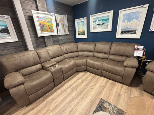 The Sapphire Corner Group, a spacious L-shaped brown recliner with cup holders, is displayed on wood flooring. Framed landscape art decorates the dark blue wall behind it, and a price tag is attached to one of the seats.