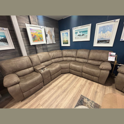 The Sapphire Corner Group, a spacious L-shaped brown recliner with cup holders, is displayed on wood flooring. Framed landscape art decorates the dark blue wall behind it, and a price tag is attached to one of the seats.
