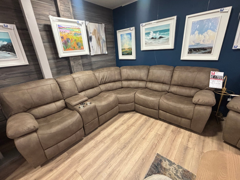 The Sapphire Corner Group, a spacious L-shaped brown recliner with cup holders, is displayed on wood flooring. Framed landscape art decorates the dark blue wall behind it, and a price tag is attached to one of the seats.