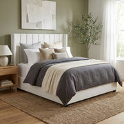 A modern bedroom with the Kildare Bedframe in beige velvet featuring a winged headboard and vertical lines, neutral bedding, a textured throw blanket, wooden nightstand with lamp, potted plant, abstract wall art, sheer curtains, and a woven rug on wood floors.