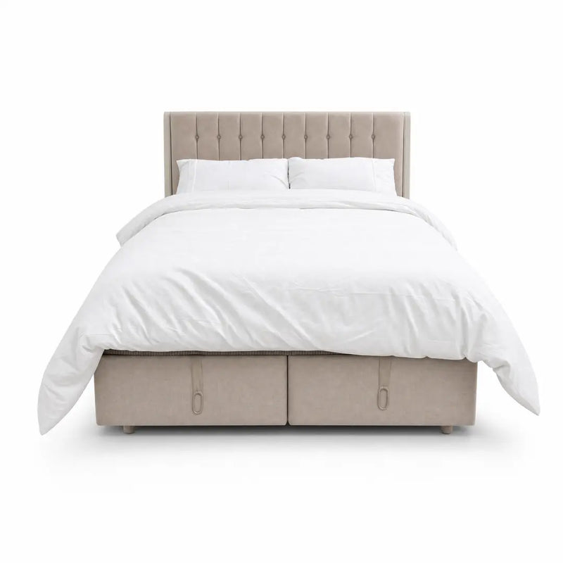 The Bella Ottoman Bed, featuring beige upholstery, a tall tufted headboard, and ample storage, is pictured with white bedding—including pillows and a duvet—against a plain white background, making it a stylish addition to your bedroom furniture collection.