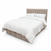 The Bella Ottoman Bed features beige upholstery, a tall tufted headboard, two base drawers for added storage, and comes styled with crisp white bedding—all showcased against a clean white backdrop for a sleek, contemporary look.