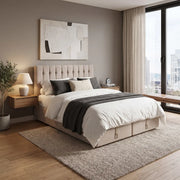 Modern bedroom featuring the Bella Ottoman Bed, a beige upholstered bed offering extra storage, adorned with white bedding and a gray throw blanket. A nightstand with lamp sits beside the bed, while abstract wall art hangs above. The space includes a large window with city views, beige curtains, a potted plant, and a woven rug on wooden floors. The sleek design of the Bella Ottoman Bed perfectly complements the contemporary aesthetic.