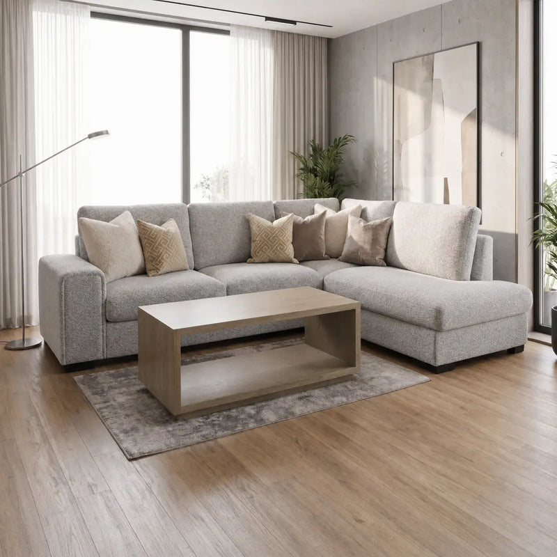 A modern living room featuring the Sorrento Chaise Corner sectional sofa in a light gray neutral textured fabric, complemented by beige and patterned pillows, a wooden coffee table, a floor lamp, potted plants, large windows with sheer curtains, and sleek wooden floors.