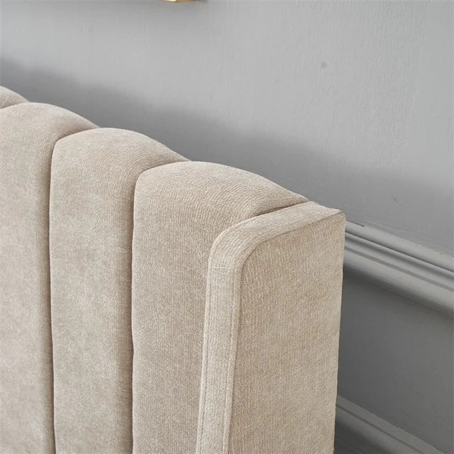 A close-up of the Oscar Bedframe showcases its beige upholstery and vertical, channel-tufted headboard, set against a gray wall with decorative molding for a modern bedroom look.