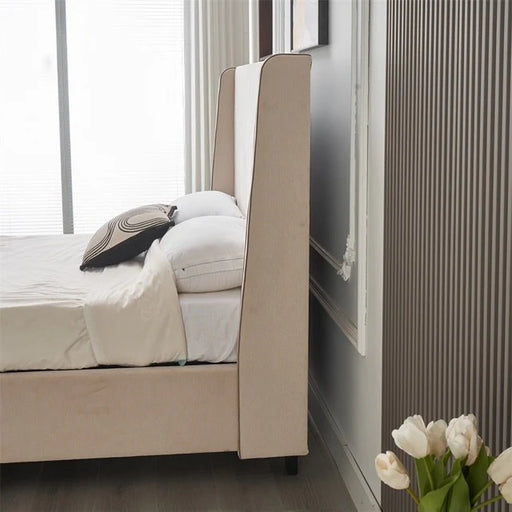 A modern bedroom featuring the Rivera Storage Bedframe in beige linen, white bedding, and a geometric pillow. White tulips in a vase rest by ribbed wall paneling and a large window with blinds in the background.