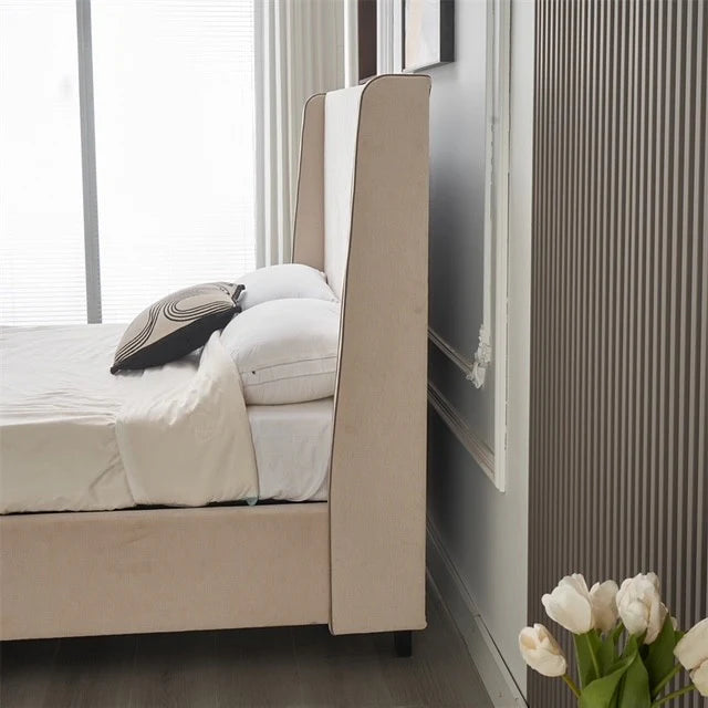 A modern bedroom featuring the Rivera Storage Bedframe in beige linen, white bedding, and a geometric pillow. White tulips in a vase rest by ribbed wall paneling and a large window with blinds in the background.