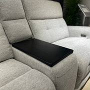A close-up of the Avery Electric Recliner Corner shows its modern light gray fabric upholstery, with a black rectangular tray placed over the center seat. In the background, a green plant and a stand with a sign are visible.