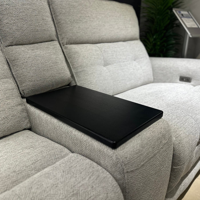 A close-up of the Avery Electric Recliner Corner shows its modern light gray fabric upholstery, with a black rectangular tray placed over the center seat. In the background, a green plant and a stand with a sign are visible.