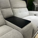 A close-up of the Avery Electric Recliner Corner shows its modern light gray fabric upholstery, with a black rectangular tray placed over the center seat. In the background, a green plant and a stand with a sign are visible.
