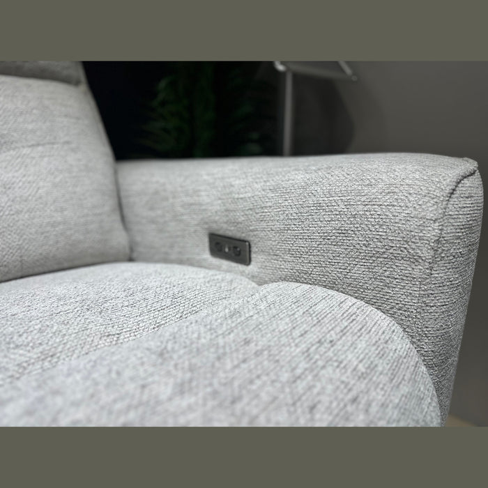 Close-up of the Avery Electric Recliner Corner in light gray fabric, featuring a built-in USB port and control buttons on the armrest—an ideal addition to a modern living room sofa arrangement. A blurred dark green plant appears in the background.