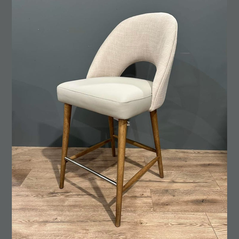 The Florence Counter Stool features a curved light beige upholstered backrest and seat, wooden legs, and a metal footrest, shown on a light wood floor against a gray wall.