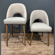Two Florence Counter Stools with light fabric seats and wooden legs are placed side by side on a wooden floor against a gray wall. The left stool is upholstered, while the right one has a faux leather look and is slightly shorter.