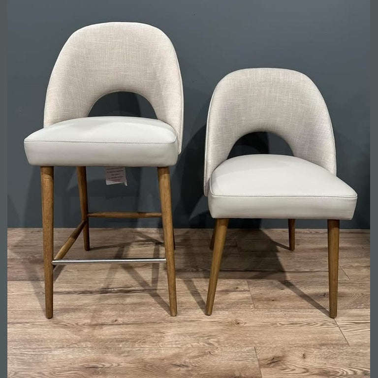 Two Florence Counter Stools with light fabric seats and wooden legs are placed side by side on a wooden floor against a gray wall. The left stool is upholstered, while the right one has a faux leather look and is slightly shorter.