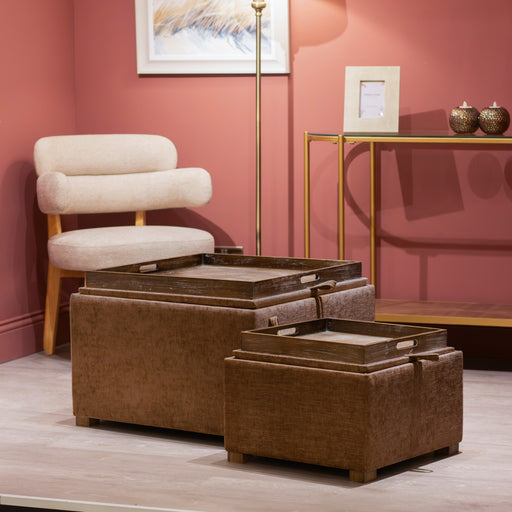 A cozy room with a beige chenille fabric chair, a gold-framed console table, and Maya tray table pouffes in Cappuccino (set of 2) with hidden storage trays on top, all set against a muted pink wall decorated with art.