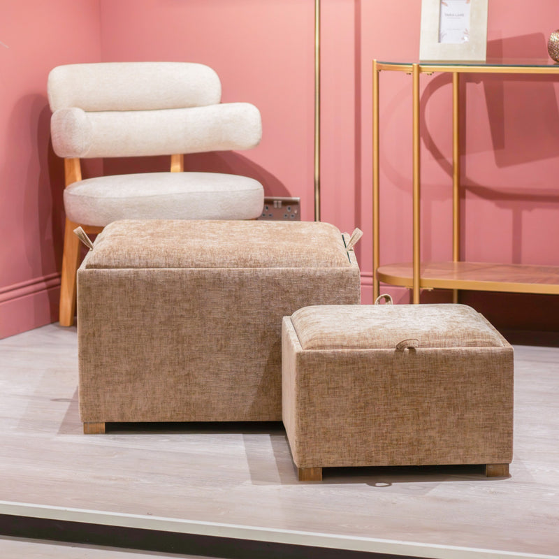 A modern interior scene featuring a cream upholstered chair, a gold metal and wood shelving unit, and the Maya tray table pouffes in Cappuccino (set of 2) on a light wood floor against a pink wall.