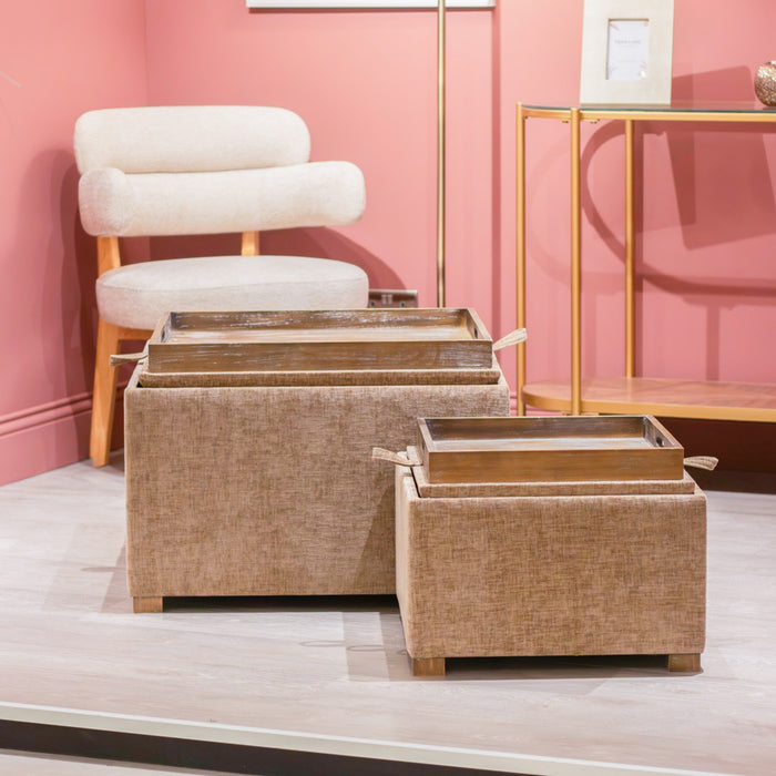 The Maya tray table pouffes in Cappuccino (set of 2) feature hidden storage and wooden trays, placed on a light wood floor. Behind them, a cream armchair and gold console table stand against a pink wall.