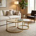 A modern living room featuring the Salzburg Nest, a set of gold-framed marble coffee tables, arranged beside a beige sofa, with a floor lamp, large potted plant, and a dark accent chair by the window atop a textured rug.