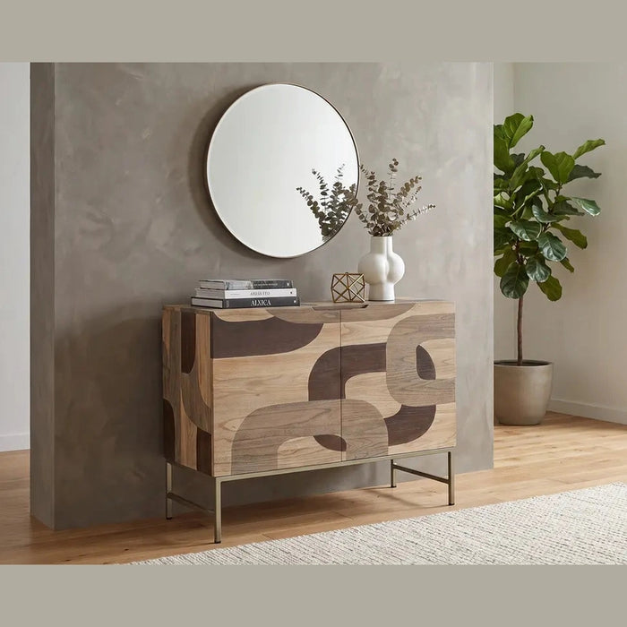 The Mosaic 2 Door Sideboard with a natural wood finish and abstract geometric pattern stands against a gray wall, adorned with decor and books. Above it hangs a round mirror. To the right, there is a potted plant near a large window, while a rug covers the wooden floor.