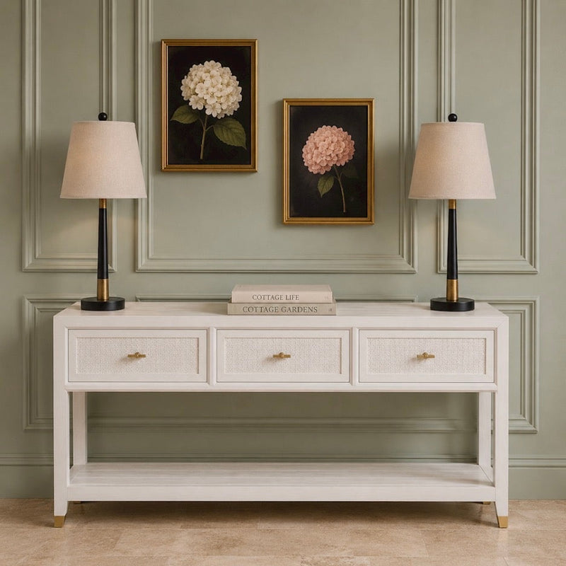 A coastal-inspired Bali 3 Drawer Console, offering generous storage with its three drawers, is styled with two beige lamps, a stack of books in the center, and two framed hydrangea paintings displayed on a light green paneled wall.