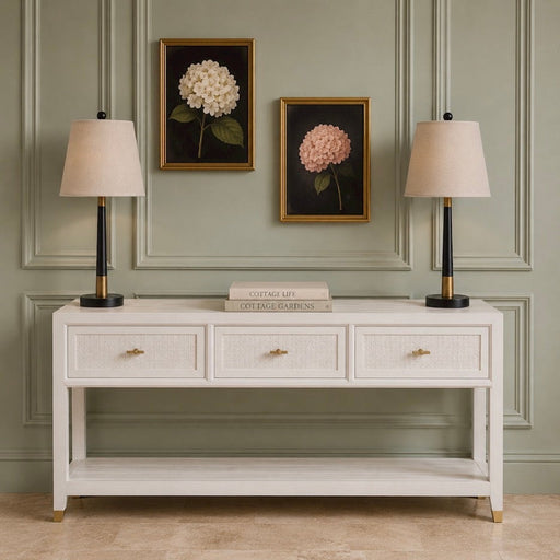 A coastal-inspired Bali 3 Drawer Console, offering generous storage with its three drawers, is styled with two beige lamps, a stack of books in the center, and two framed hydrangea paintings displayed on a light green paneled wall.