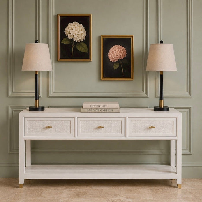 A coastal-inspired Bali 3 Drawer Console, offering generous storage with its three drawers, is styled with two beige lamps, a stack of books in the center, and two framed hydrangea paintings displayed on a light green paneled wall.