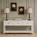 A coastal-inspired Bali 3 Drawer Console, offering generous storage with its three drawers, is styled with two beige lamps, a stack of books in the center, and two framed hydrangea paintings displayed on a light green paneled wall.