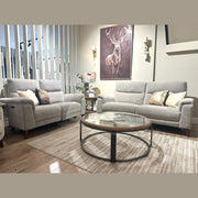 A modern living room featuring two Delta Electric Recliner sofas in grey, a round glass coffee table, a beige rug, a lamp, and a deer painting on the wall. Vertical white slats partially separate the space.