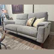 The Delta Electric Recliner, a modern light gray sofa with wooden legs, two accent pillows, and a beige and gold cushion, sits on a striped rug next to a glass coffee table and lamp in a cozy living room setting, offering modern comfort and easy-clean fabric for your space.