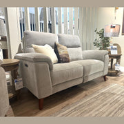A light gray Delta Electric Recliner with two pillows sits on a rug in a living room display, next to a round wooden side table and a lit lamp, with price tags attached to the furniture.
