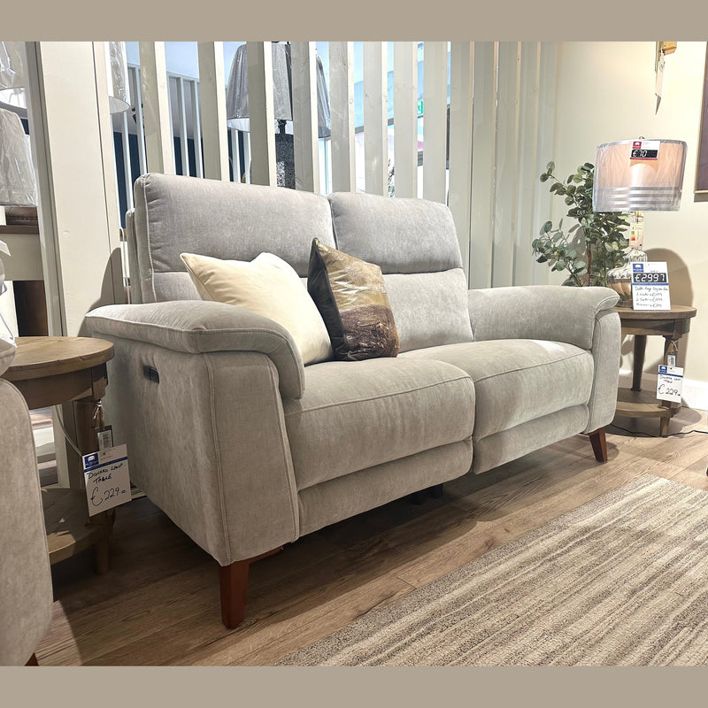 A light gray Delta Electric Recliner with two pillows sits on a rug in a living room display, next to a round wooden side table and a lit lamp, with price tags attached to the furniture.
